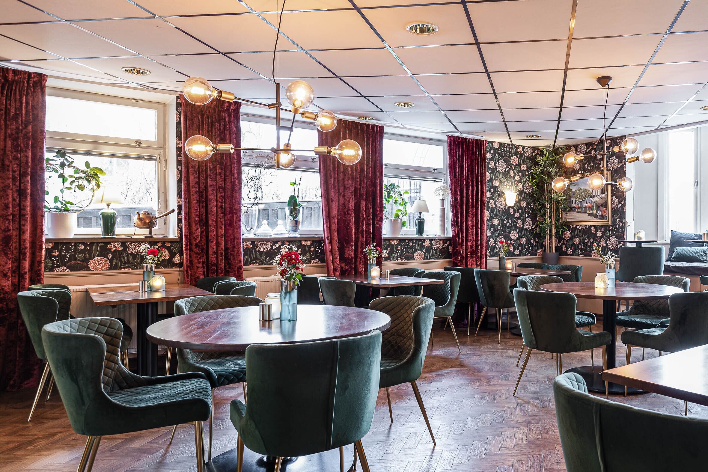 Un restaurant avec des tables rondes et des chaises à revêtement vert, doté d’un plafond carrelé et de rideaux rouges.