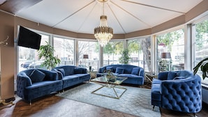 A spacious living room with a large chandelier, two blue sofas, a glass coffee table, and a view of the city through large windows. - Hotell Arkad (Vasteras)