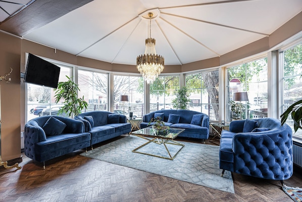 A spacious living room with a large chandelier, two blue sofas, a glass coffee table, and a view of the city through large windows.