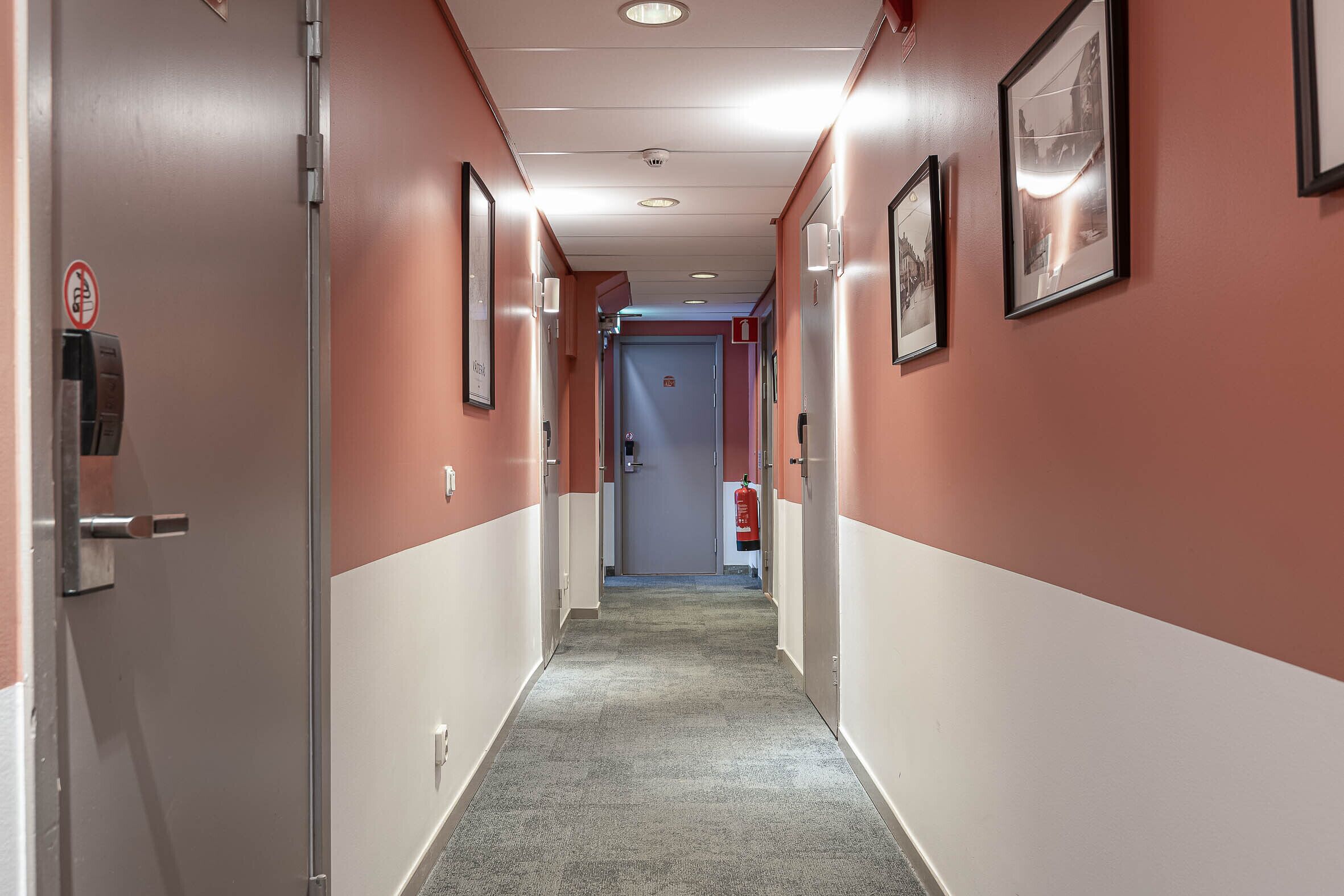 A corridor with a fire extinguisher, a no-smoking sign, and framed pictures on the walls.