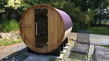Une structure de sauna en bois, dotée d’une porte, entourée d’un jardin avec un banc et une clôture en treillis.