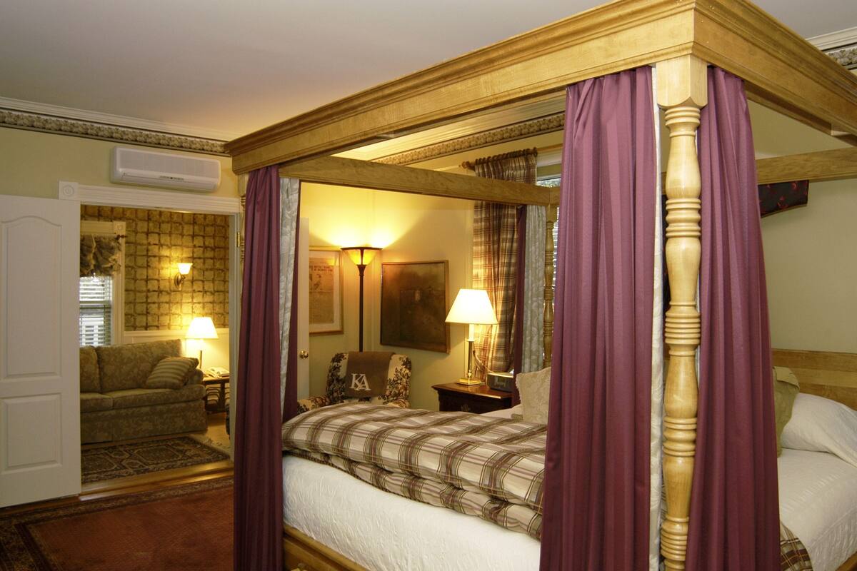 Un lit à baldaquin avec une tête de lit, une table de chevet avec une lampe, un canapé et un tapis dans une chambre d’hôtel.