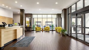 Un vestíbulo de hotel con mostrador de recepción, sillones amarillos, plantas en macetas y ventanas amplias.