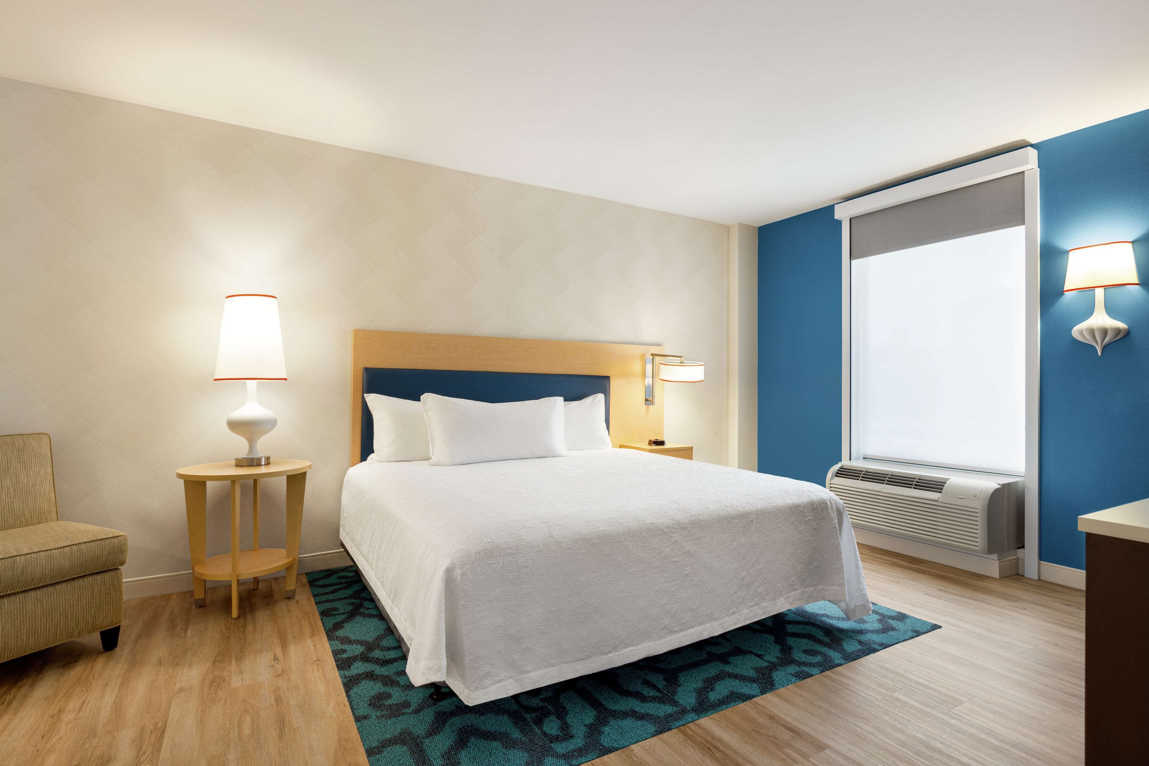A hotel room with a bed, bedside tables, a chair, a window with blinds, and an air conditioning unit.