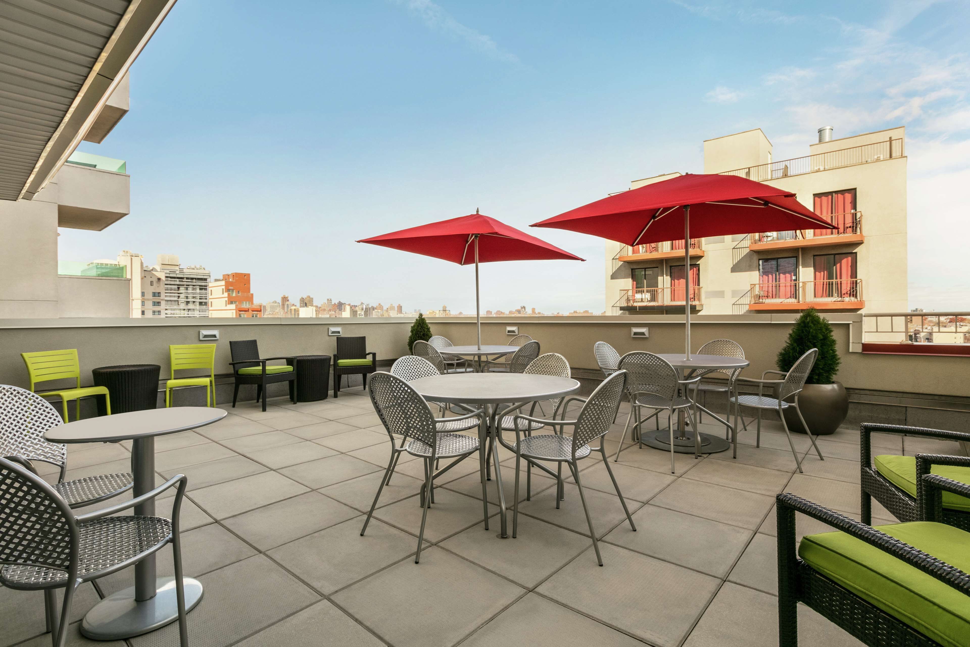 A rooftop terrace with outdoor seating, including round tables and chairs, and red umbrellas providing shade.