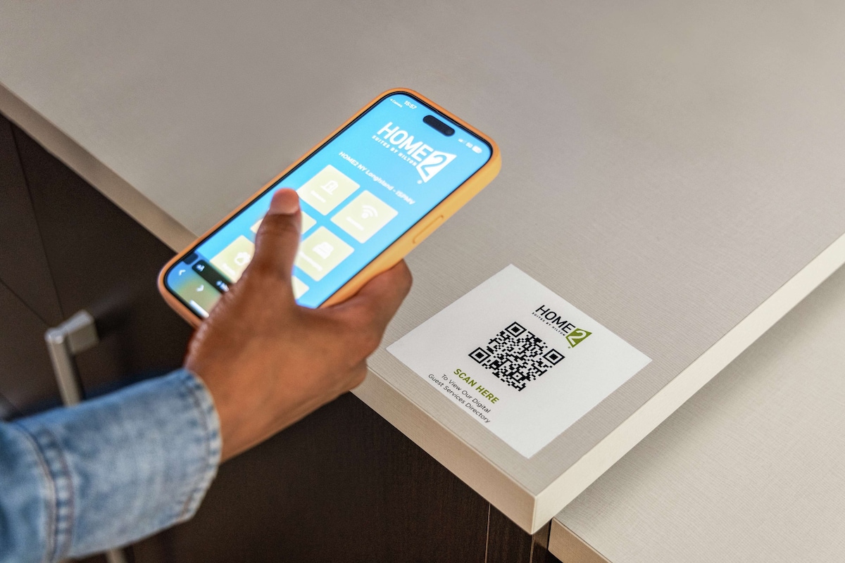 Una mano sosteniendo un teléfono inteligente con una aplicación de hotel, un código QR y un cartel que indica un directorio digital de servicios para huéspedes.