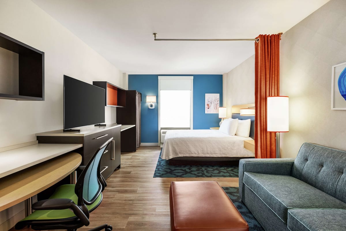 Una habitación de hotel con una cama, un escritorio con computadora, un televisor, un sofá y una ventana con cortinas.