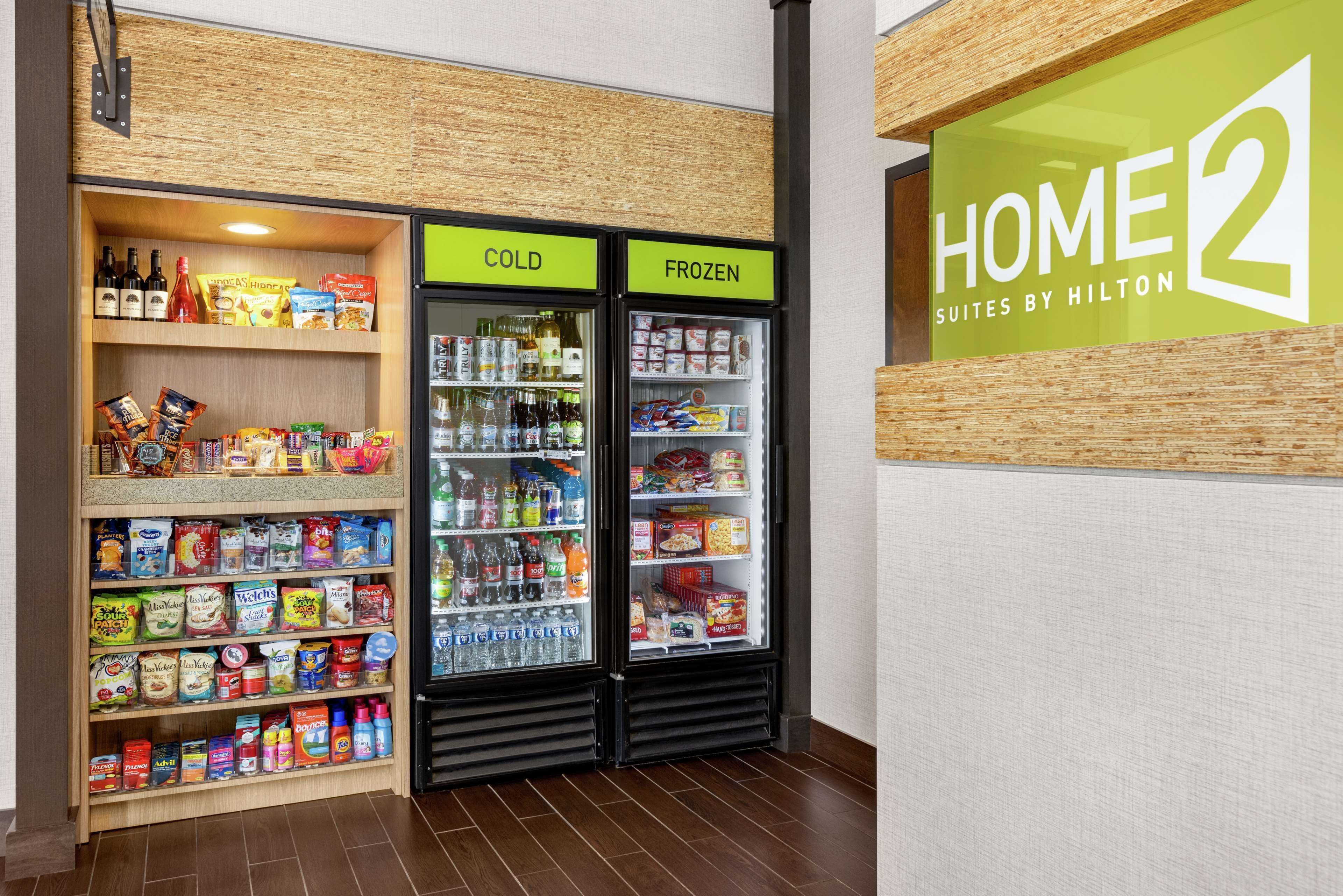 A hotel kitchenette with a cold and frozen section, shelves stocked with snacks and beverages, and a sign for 'HOME 2 Suites by Hilton'.