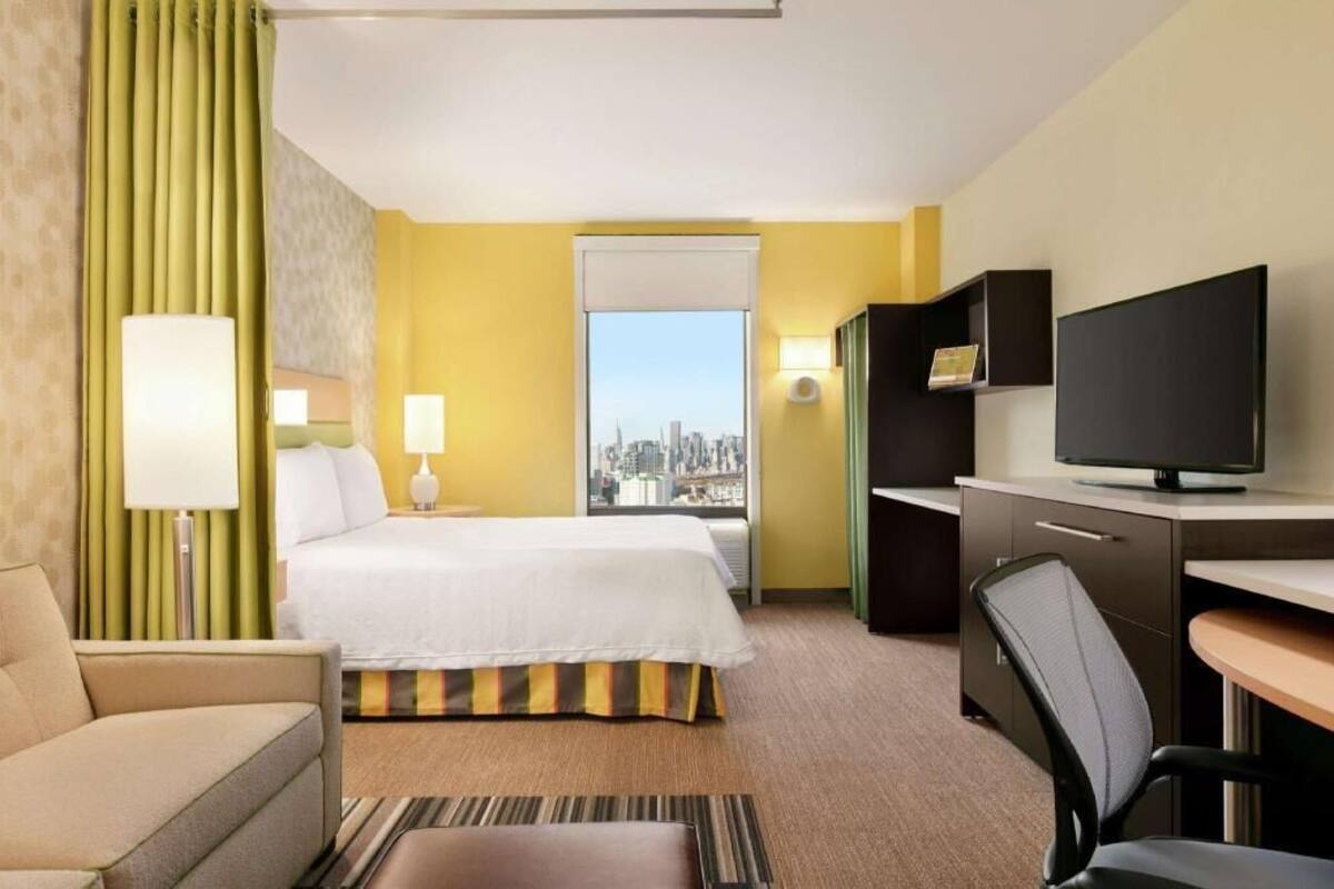 Habitación de hotel con cama, escritorio con computadora, silla, televisor y vista a la ciudad.