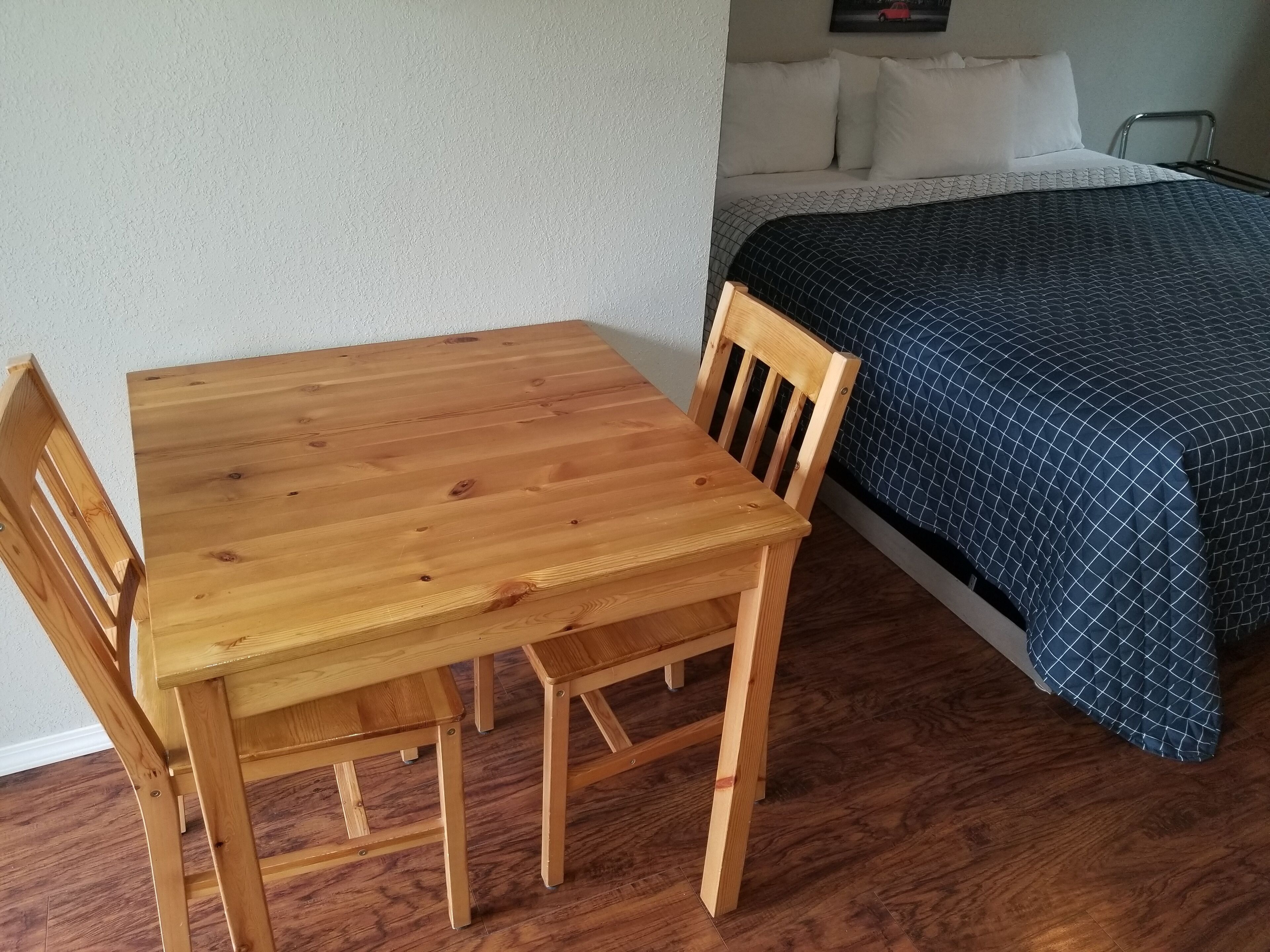 A room with a wooden table and chairs, a bed with a blue patterned bedspread, and a wooden floor.