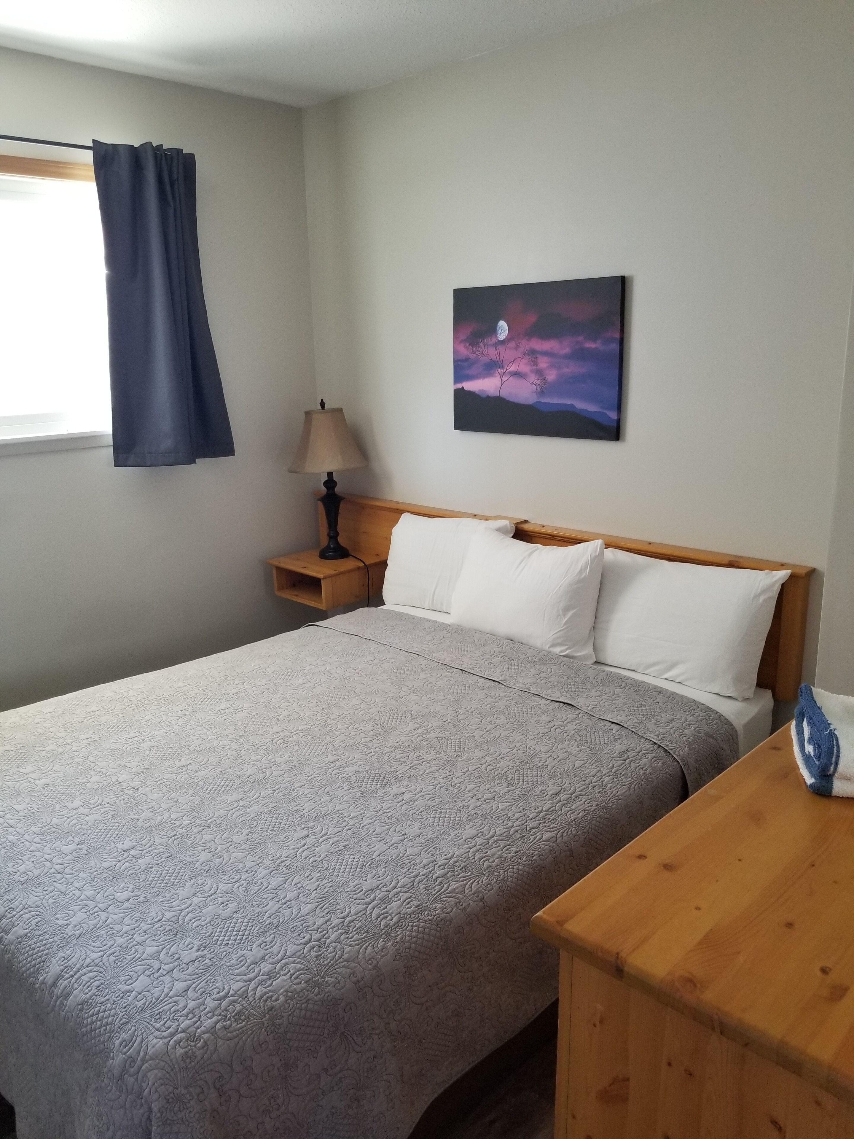 Ein ordentlich gemachtes Bett mit einem gemusterten Überwurf, einer Nachttischlampe und einem Gemälde einer mondbeschienenen Landschaft an der Wand.