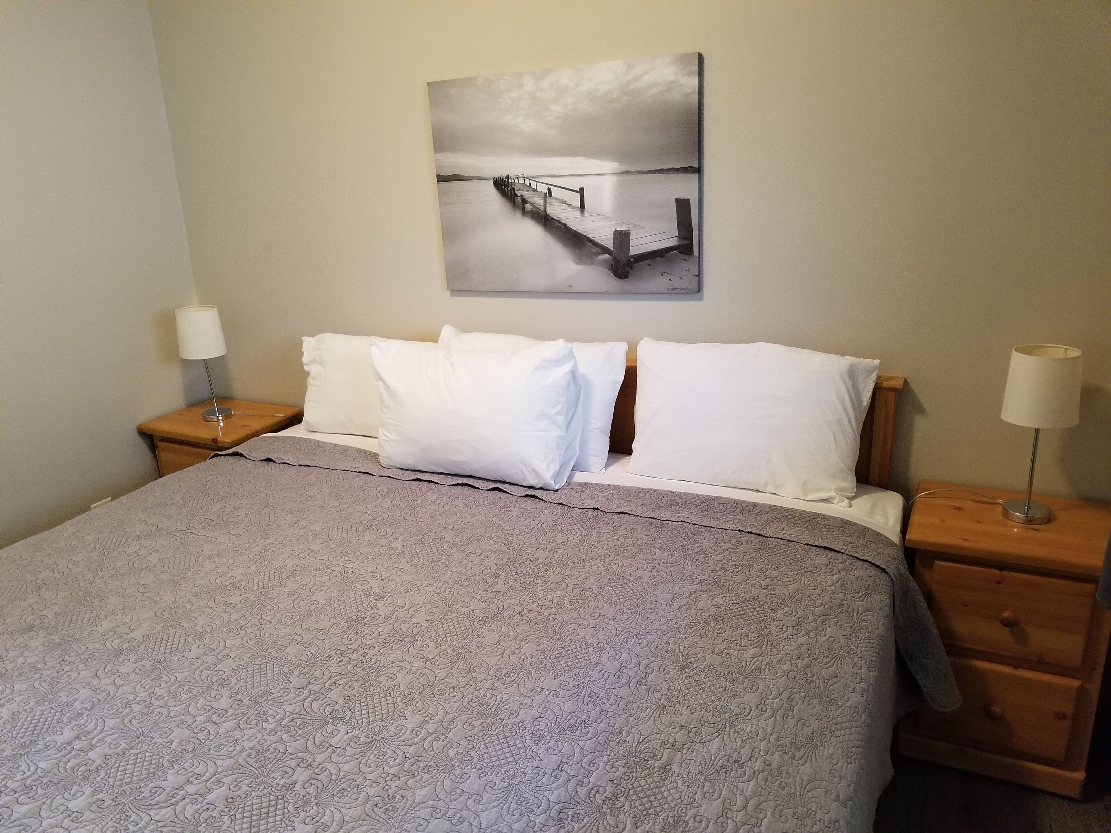 Una cama bien hecha con una colcha estampada, dos lámparas de noche y un cuadro de un muelle.