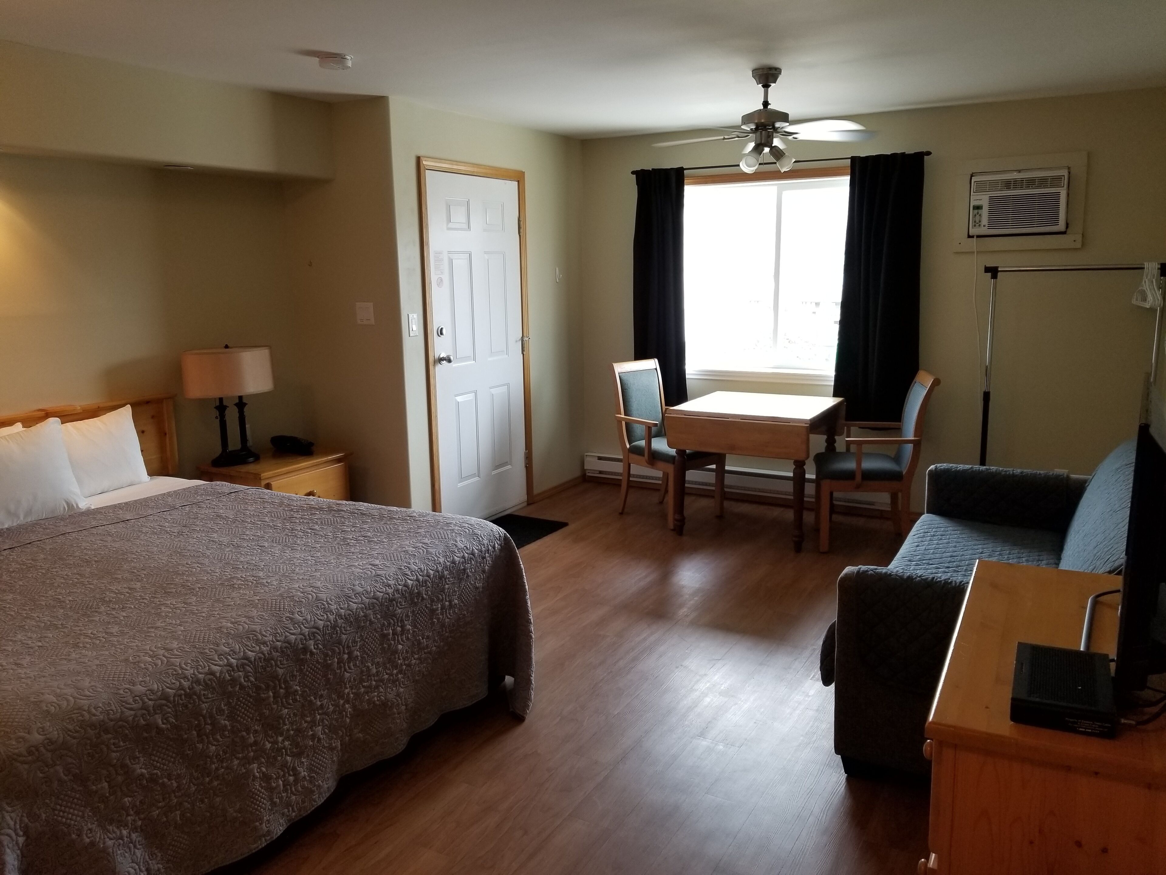 A hotel room with a bed, a desk, a chair, a sofa, a ceiling fan, and a window with curtains.