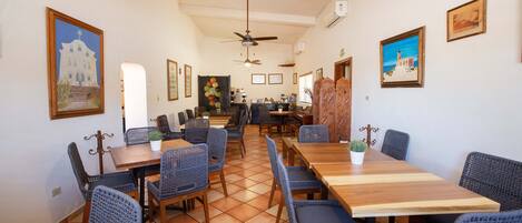 A dining area with wooden tables and blue wicker chairs, a ceiling fan, and framed artwork on the walls.