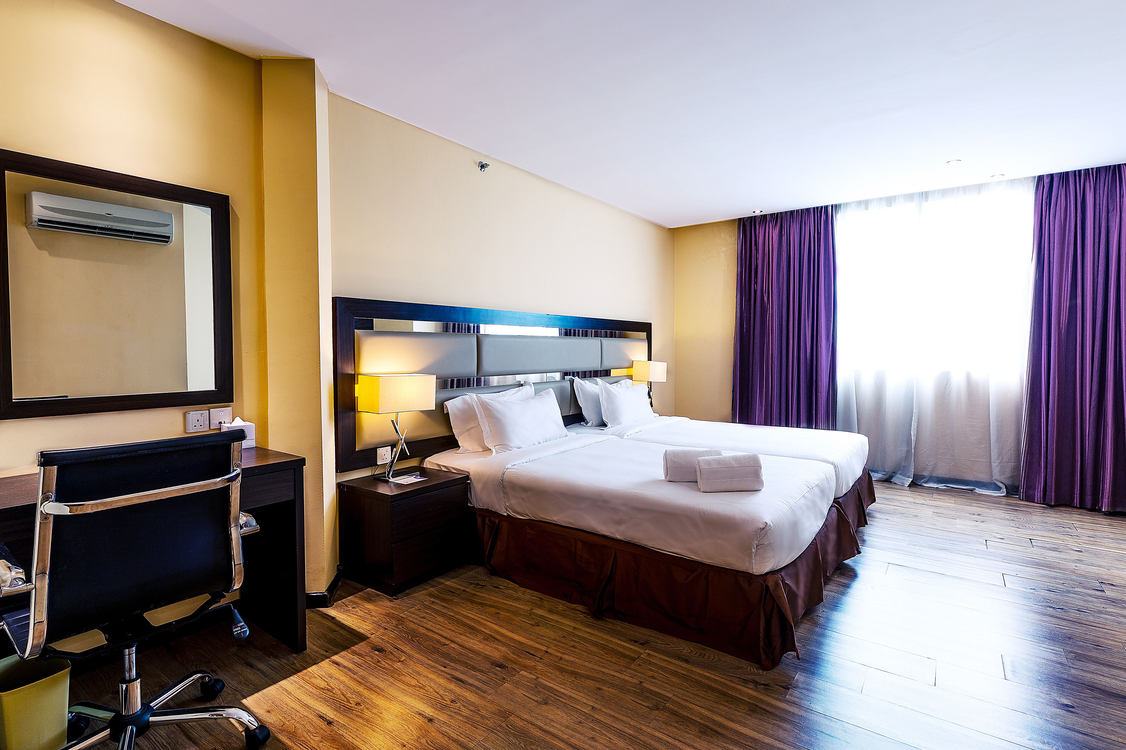 A hotel room with a large bed, a desk with a chair, a nightstand with lamps, and a window with curtains.