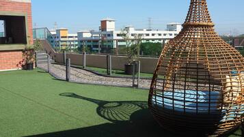 A rooftop terrace with artificial grass, a wicker chair, and a view of buildings in the background.