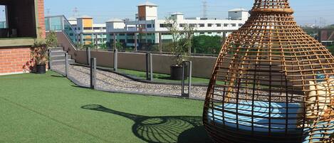 Una terraza en la azotea con césped artificial, una silla de mimbre y vistas a edificios al fondo.