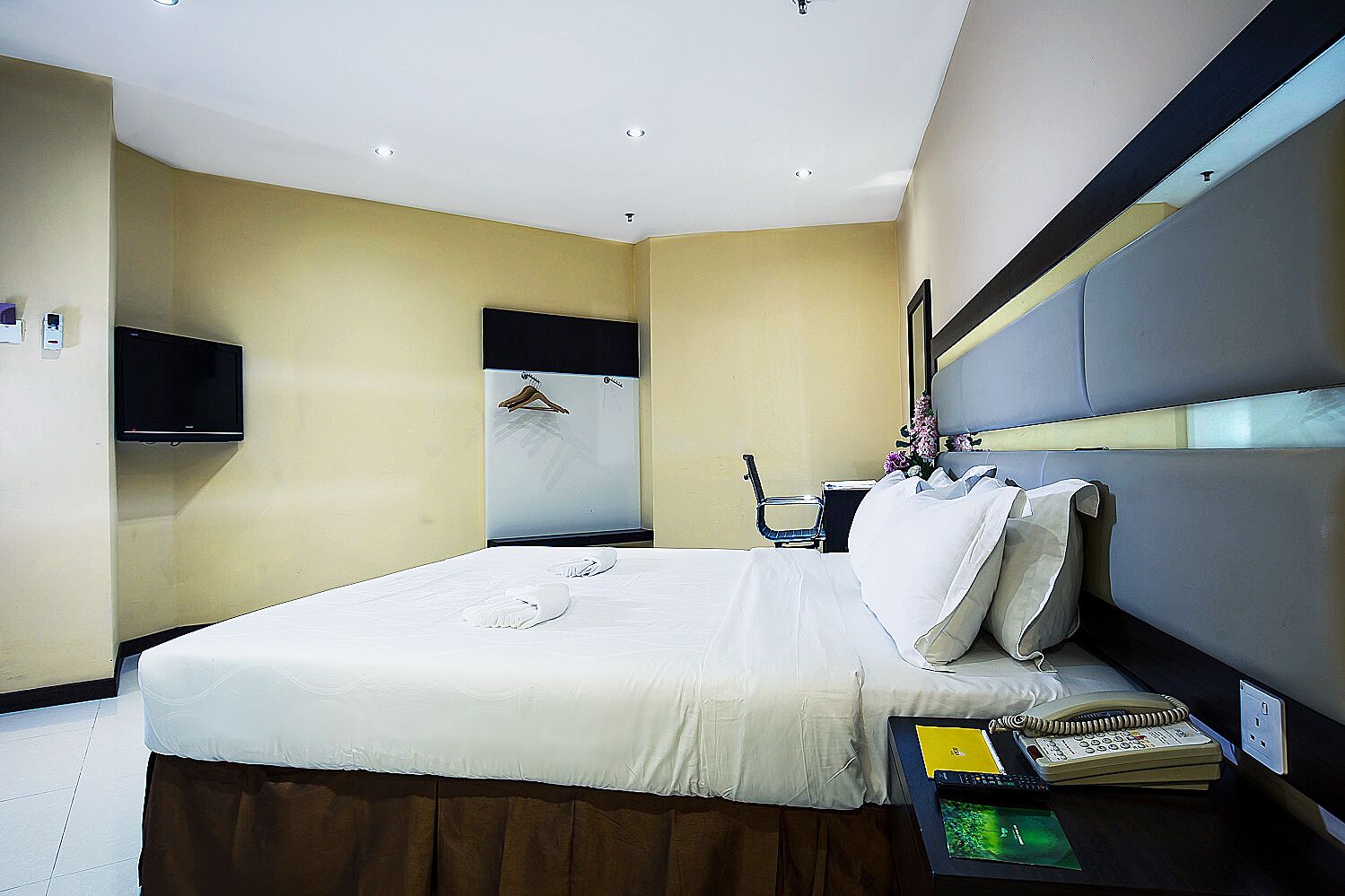 A modern hotel room with a large bed, a flat-screen TV mounted on the wall, a desk with a telephone and a notepad, and a mirror above the bed.