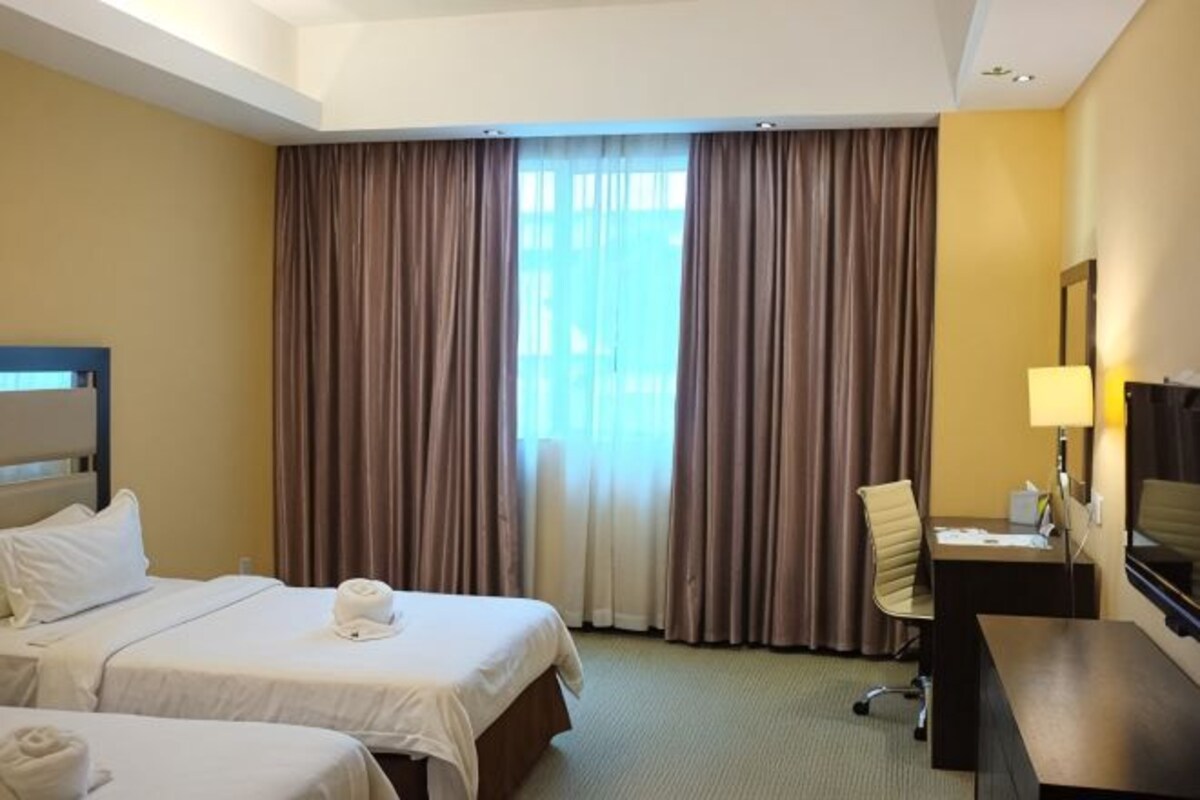 Ett hotellrum med två sängar, ett skrivbord, en stol, en TV och ett stort fönster med gardiner.