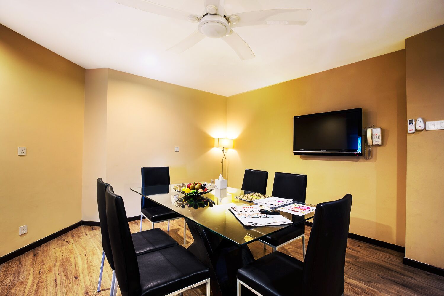 A conference room with a glass table, black chairs, a ceiling fan, a TV, and a telephone.