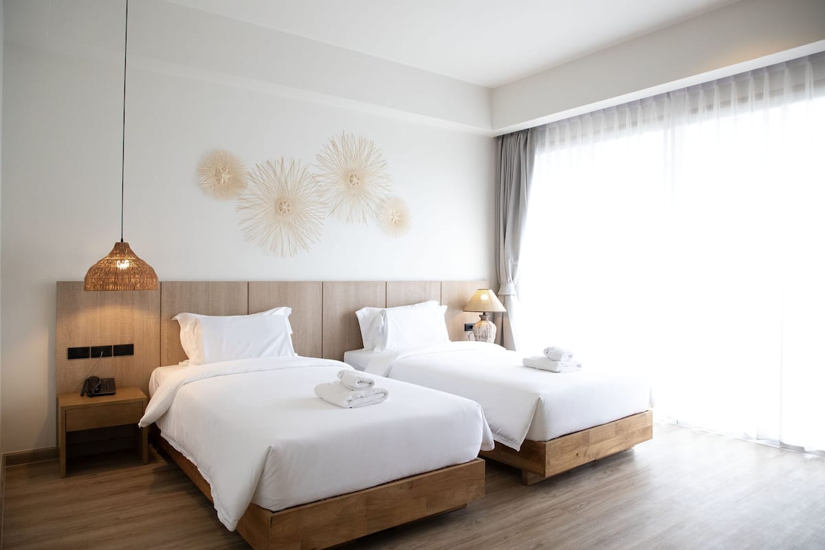 Ein Hotelzimmer mit zwei Betten, einer Hängelampe, einer Wanddekoration, einem Nachttisch und einem Fenster mit Vorhängen.