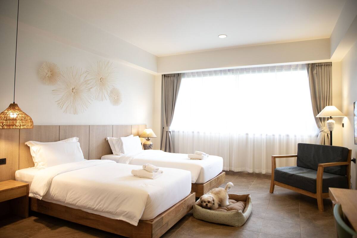 Ein Hotelzimmer mit zwei Betten, einem Holznachttisch, einem Sessel, einem Hundebett und einem großen Fenster mit Vorhängen.