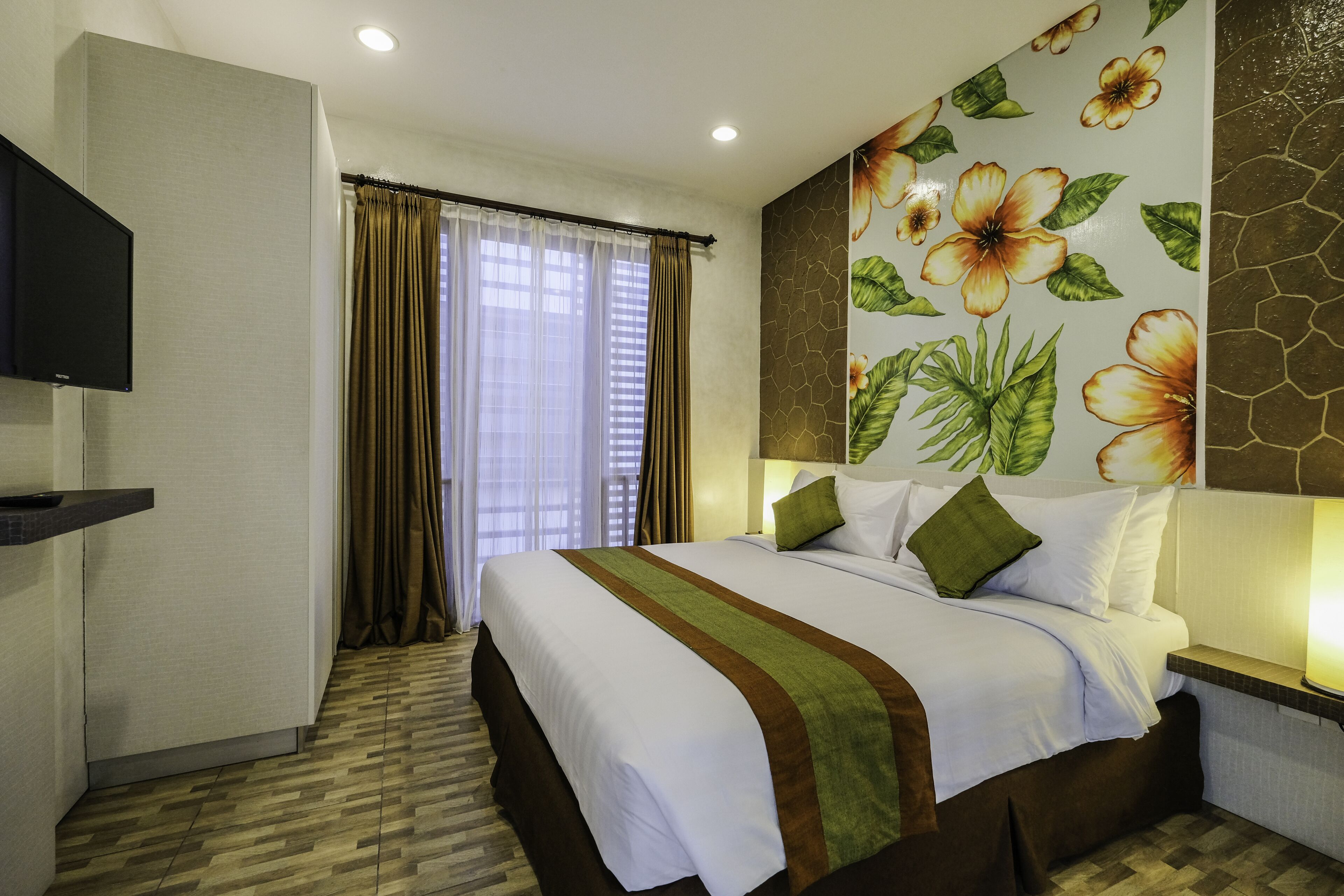 A hotel room with a large bed, a flat-screen TV, a window with curtains, and a floral wall mural.