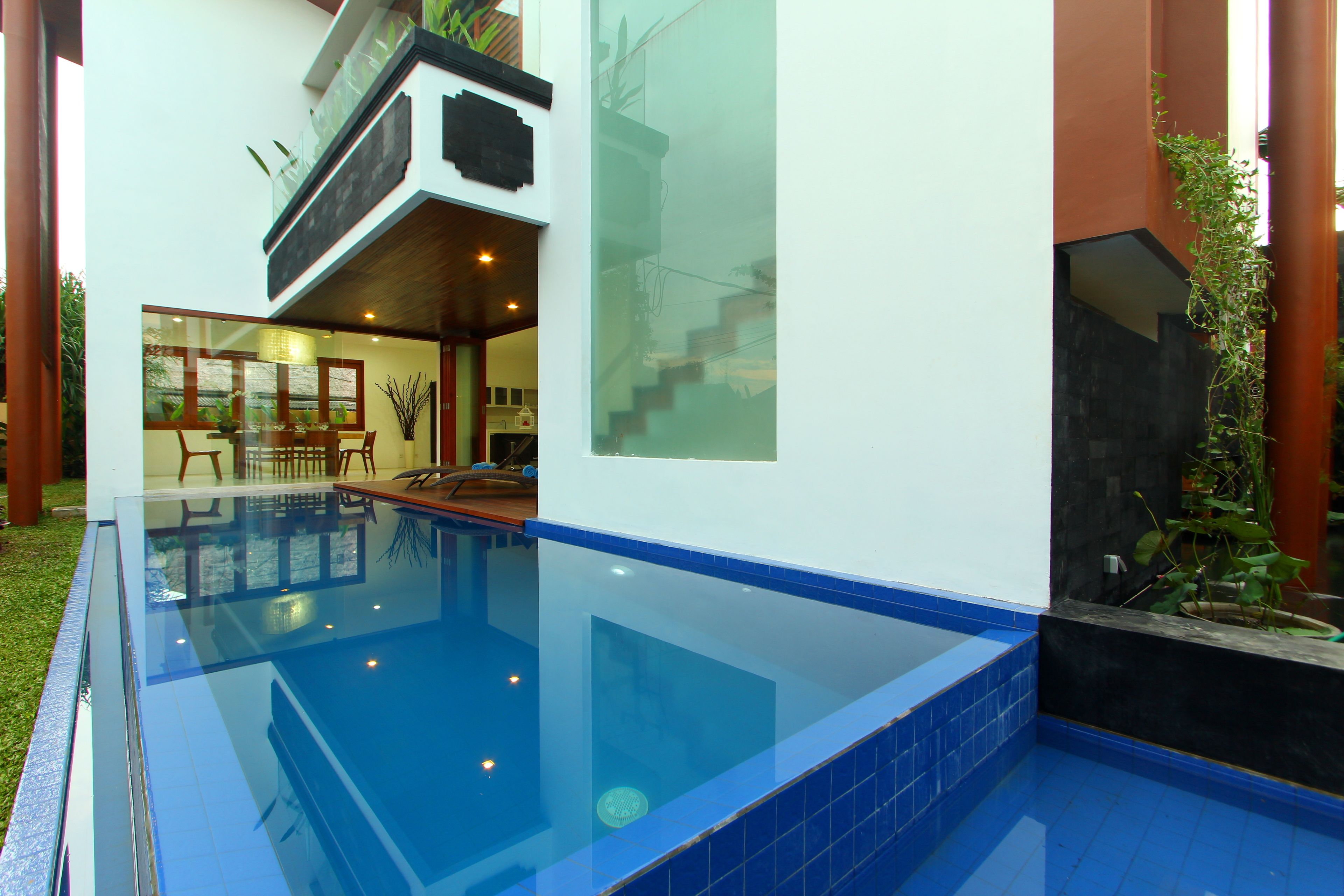 A modern indoor swimming pool with clear glass walls, a dining area with chairs and a table, and a staircase leading to an upper level.