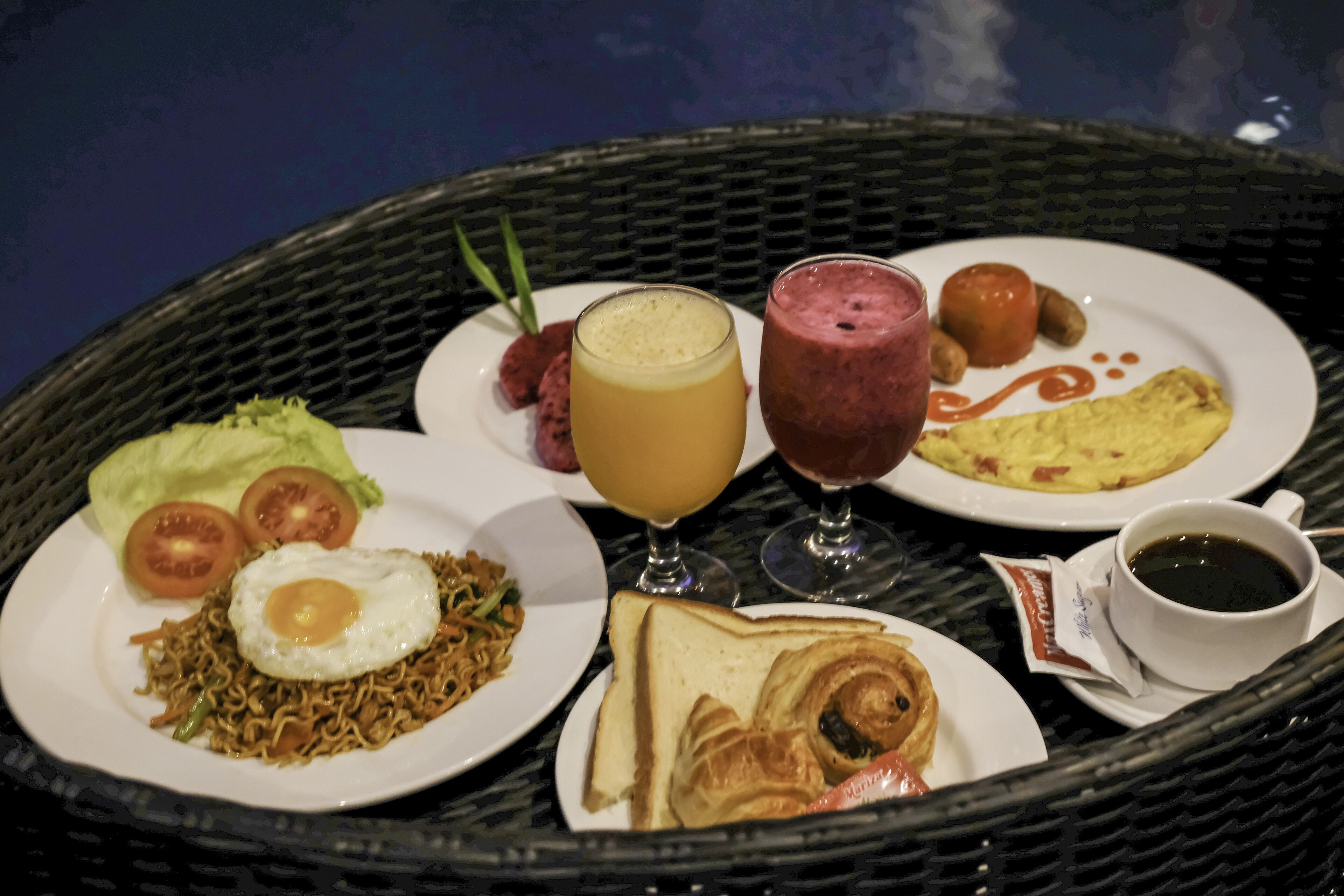 A wicker table with plates of food including noodles with an egg, toast, croissant, and a smoothie.
