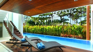 Un área de piscina interior moderna con suelo de madera, sillas de descanso y vistas a la vegetación tropical.