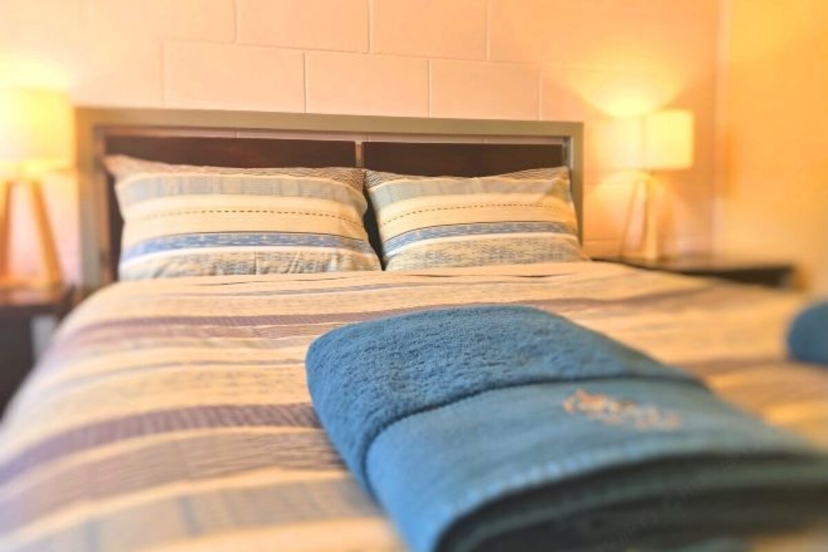 A neatly made bed with striped bedding and two pillows, a blue blanket, and a headboard against a wall with a light fixture.