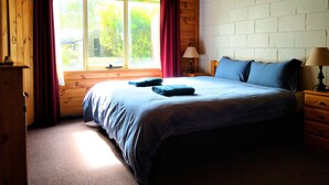 A cozy bedroom with a wooden ceiling, a large bed with blue bedding, a nightstand with a lamp, and a window with curtains. - Twelve Apostles Motel & Country Retreat (Princetown)