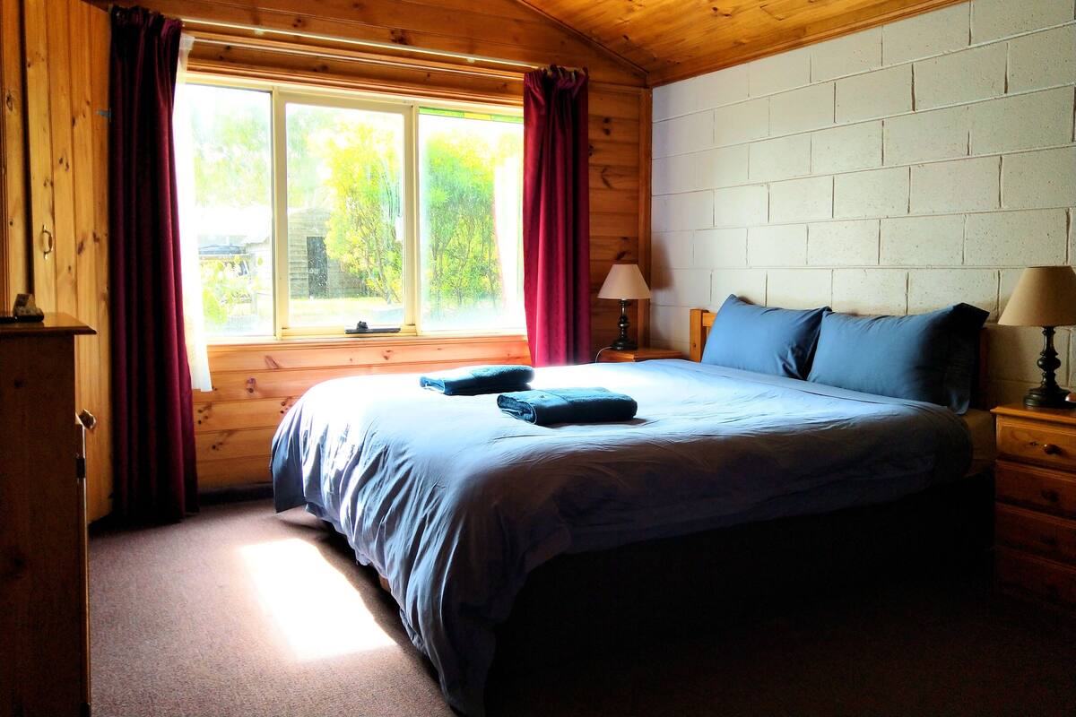 Un dormitorio acogedor con techo de madera, una cama grande con ropa de cama azul, una mesita de noche con una lámpara y una ventana con cortinas.