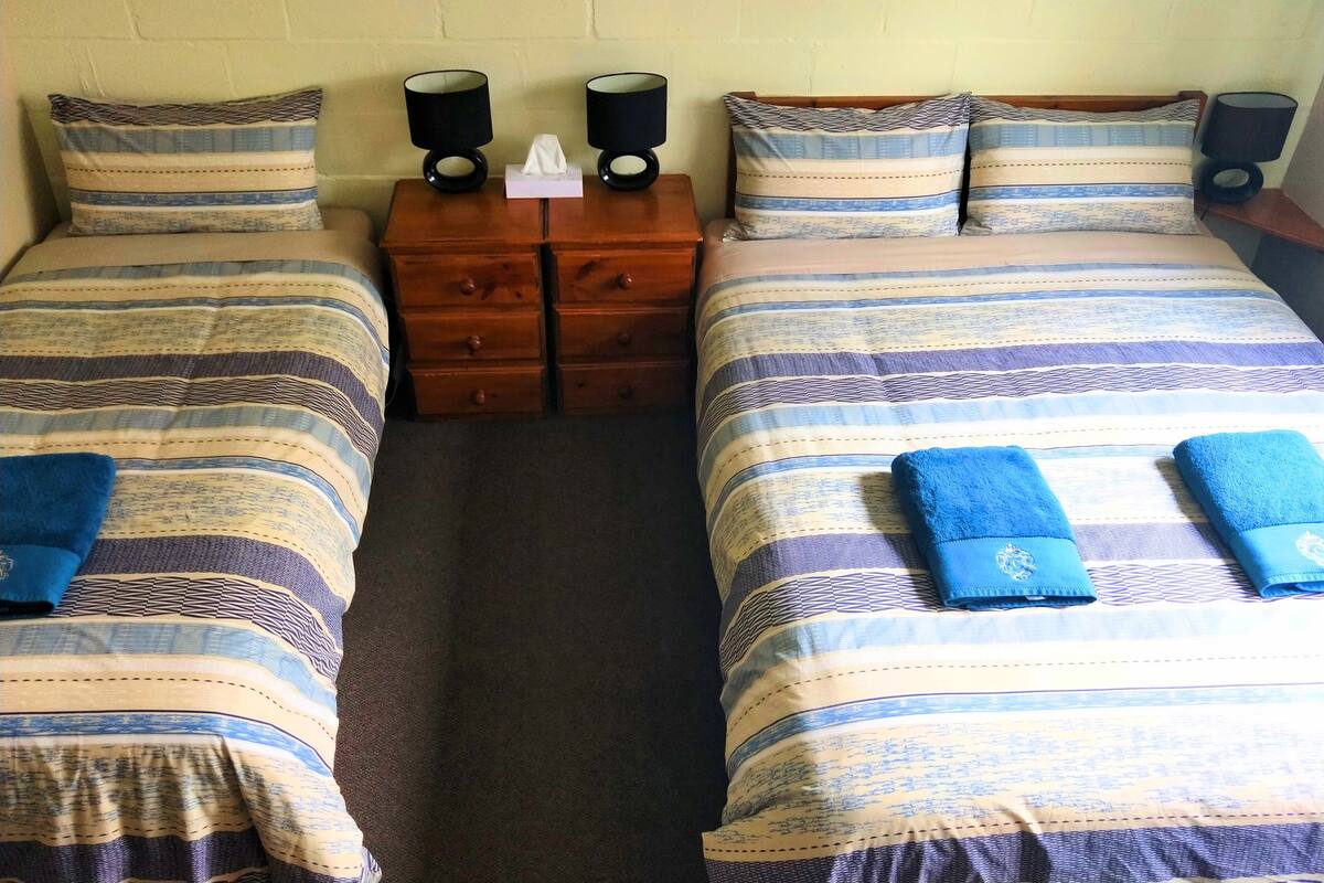 Habitación doble con dos camas, cada una con ropa de cama a rayas y toallas azules. Hay dos mesitas de noche con lámparas y una pequeña mesa con una caja de pañuelos.