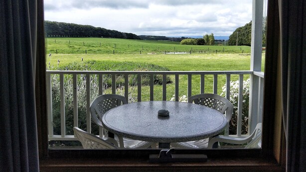 A balcony with a round table and chairs, overlooking a green field and hills. - Twelve Apostles Motel & Country Retreat (Princetown)