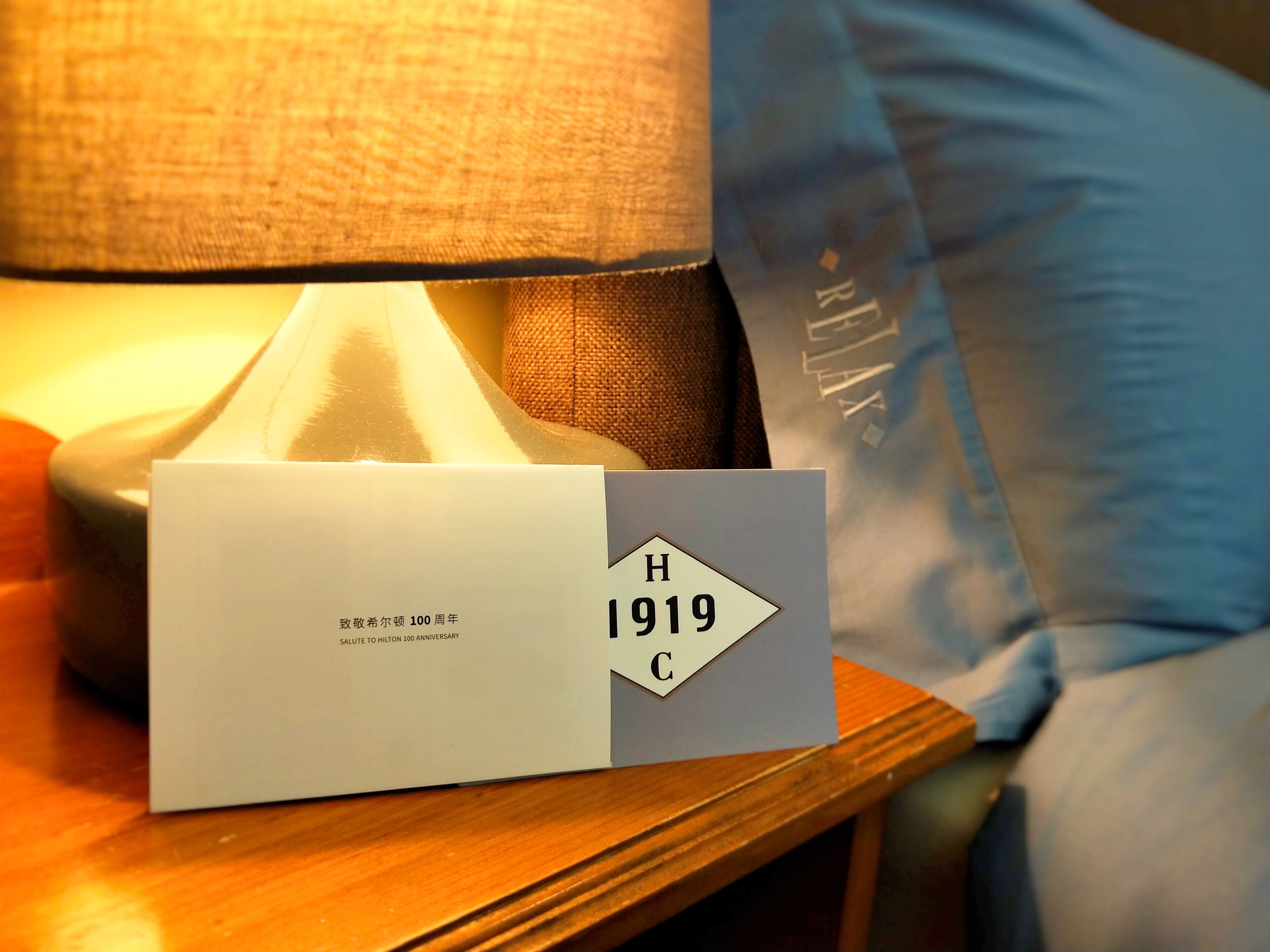 A hotel room with a bedside table, a lamp, a card with 'H 1919 C' on it, and a bed with a blue cover.