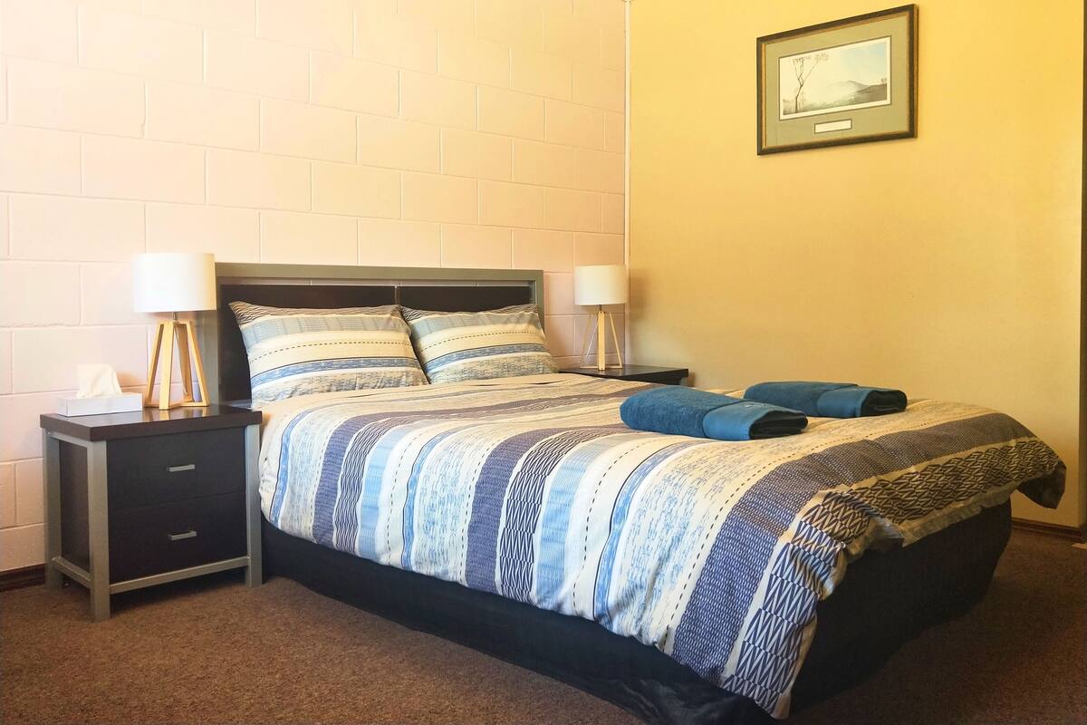 Una cama bien hecha con ropa de cama a rayas, dos lámparas de noche, una mesita de noche pequeña con una caja de pañuelos, y un cuadro en la pared.