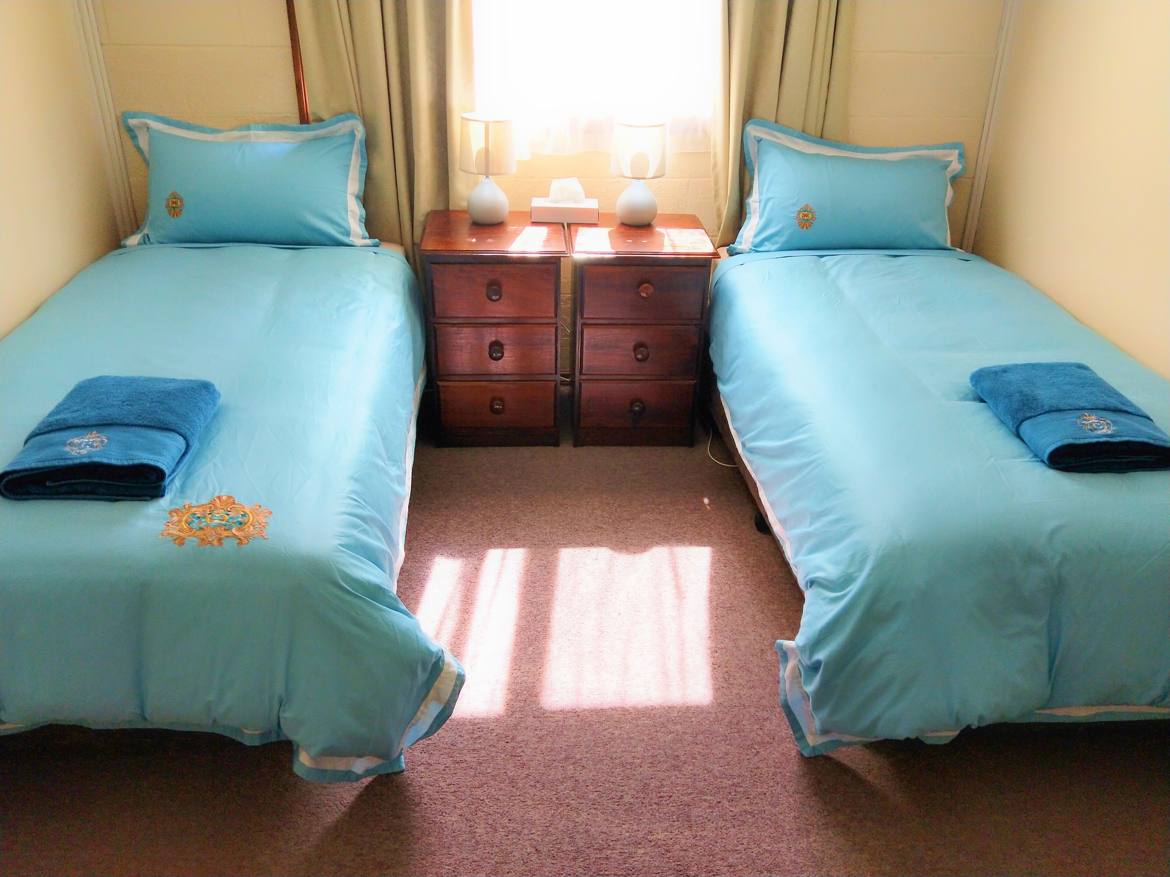 A hotel room with two single beds, each with matching blue bedding and towels, a wooden nightstand with a lamp, and a window with curtains.