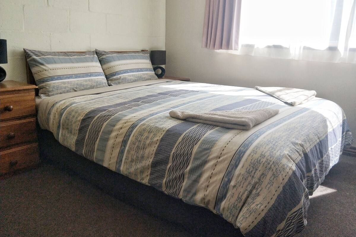 Una cama bien tendida con ropa de cama a rayas, una mesita de noche de madera con cajones, un altavoz montado en la pared y una ventana con cortinas.