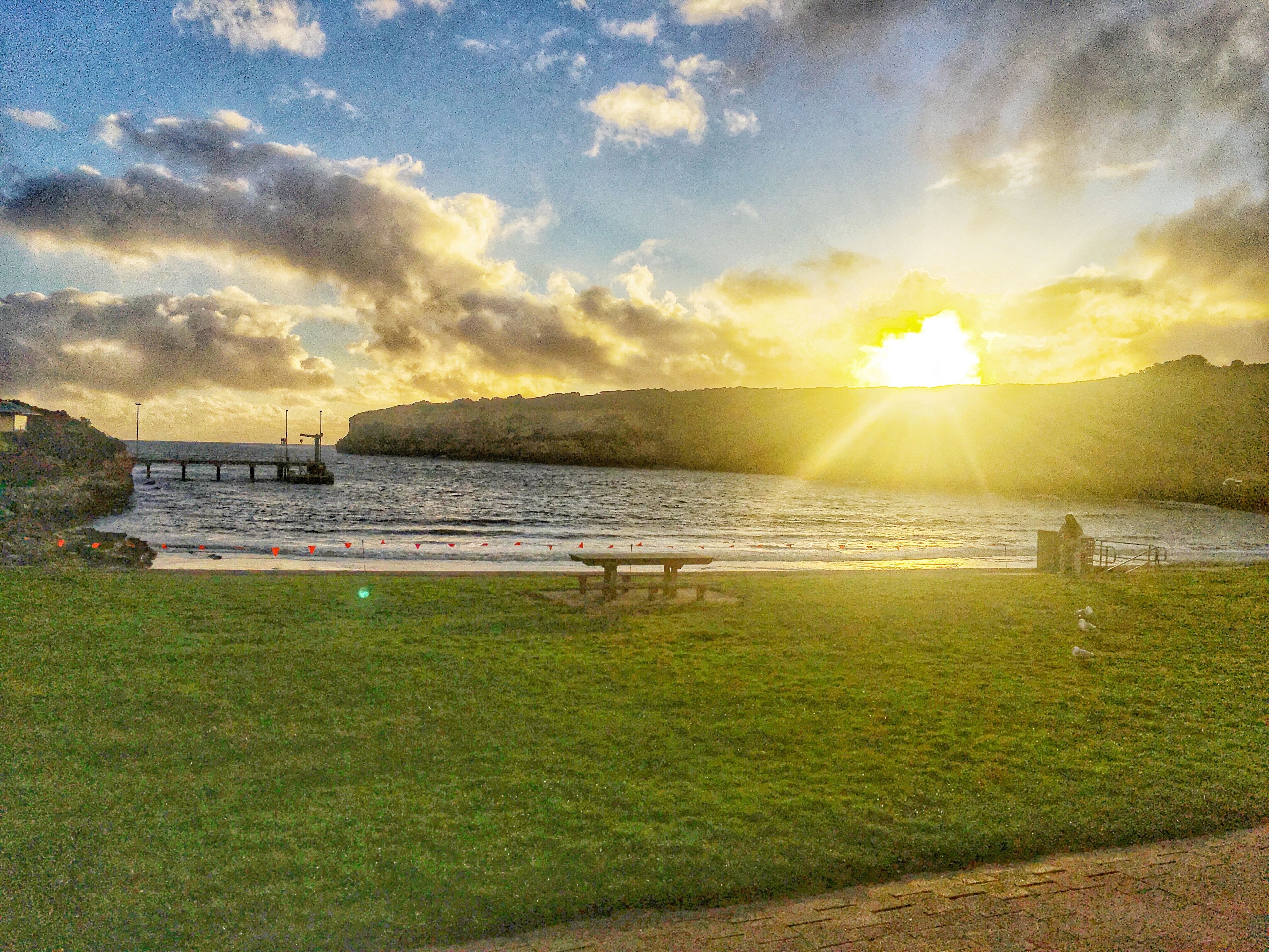 Un paisaje costero con una zona verde, un muelle y una puesta de sol sobre el agua.