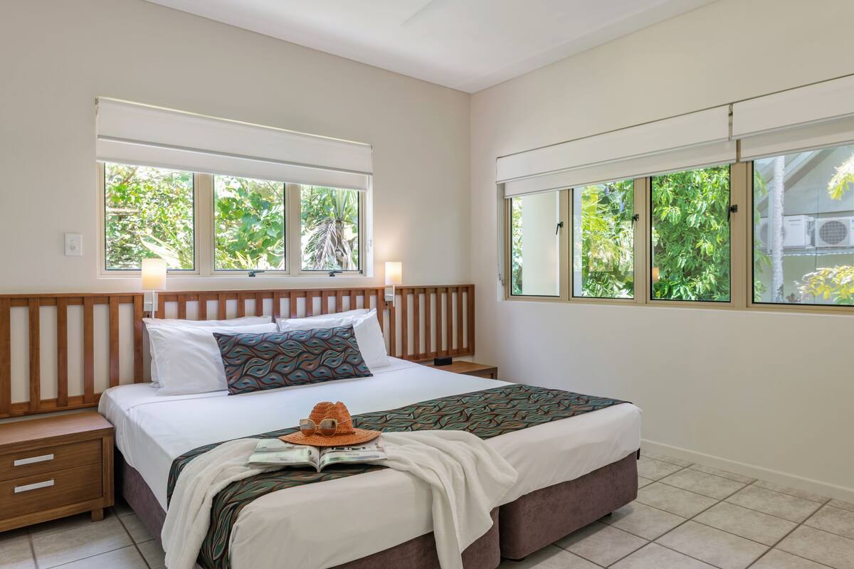 A bedroom with a wooden headboard, a large bed with white bedding, two bedside tables, and a window with a view of greenery.