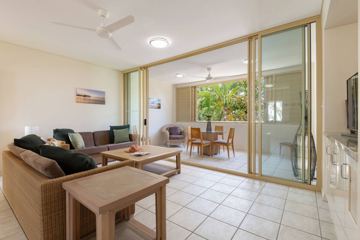 A modern living room with a sofa, coffee table, and chairs. There is a sliding glass door leading to a balcony with a view of greenery.
