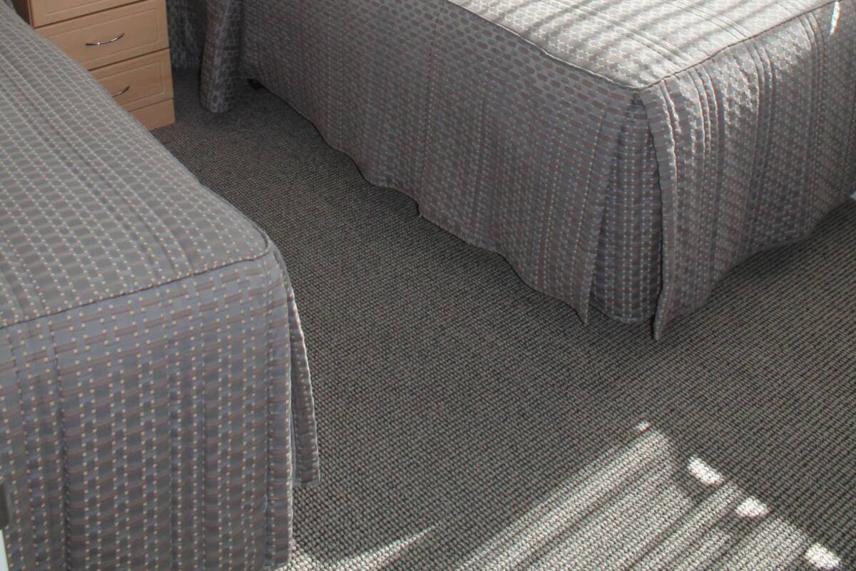 Uma cama de solteiro com colcha estampada, cômoda com gavetas, luminária na parede e uma janela com cortinas.