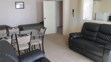 A living room with a glass dining table, chairs, a black leather sofa, and a white door leading to another room.