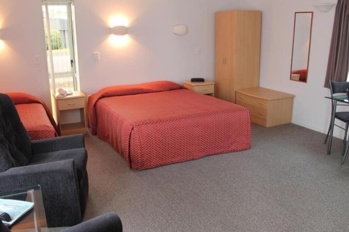 Quarto de hotel com cama, duas cadeiras, escrivaninha, guarda-roupa, espelho e janela.