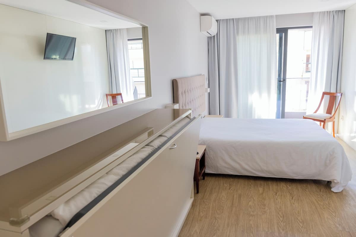 A hotel room with a bed, a bedside table, a chair, a TV mounted on the wall, and a window with curtains.