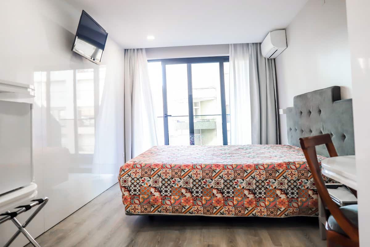 A hotel room with a bed, a TV mounted on the wall, a window with curtains, a dining table, and a chair.