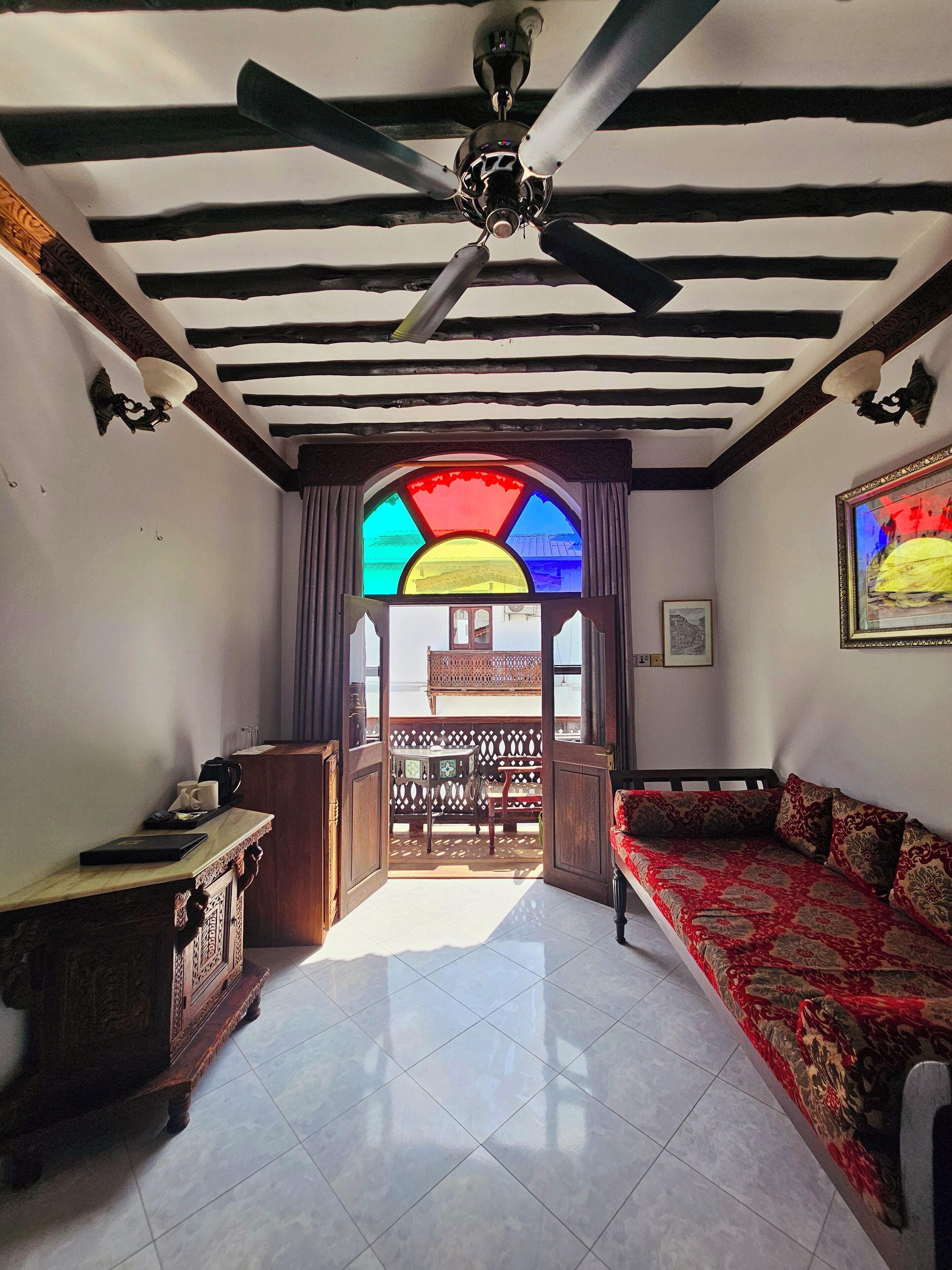A room with a ceiling fan, a sofa, a wooden cabinet, and a view through a window with a stained glass design.