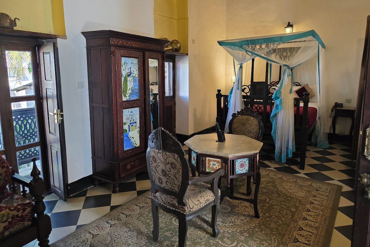 A room with a checkered floor, a wooden cabinet with glass doors, a table with chairs, and a canopy bed.