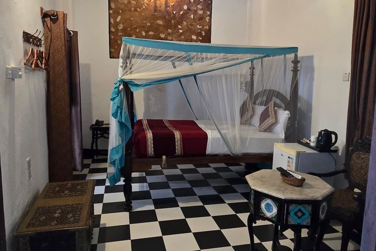A room with a checkered floor, a bed with a canopy, a small table, a chair, a wall-mounted heater, and a wall decoration.